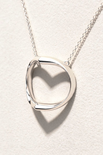 A polished sterling silver pendant necklace suspended on a delicate chain. The sculptural design casts a distinct heart-shaped shadow against a textured off-white background.