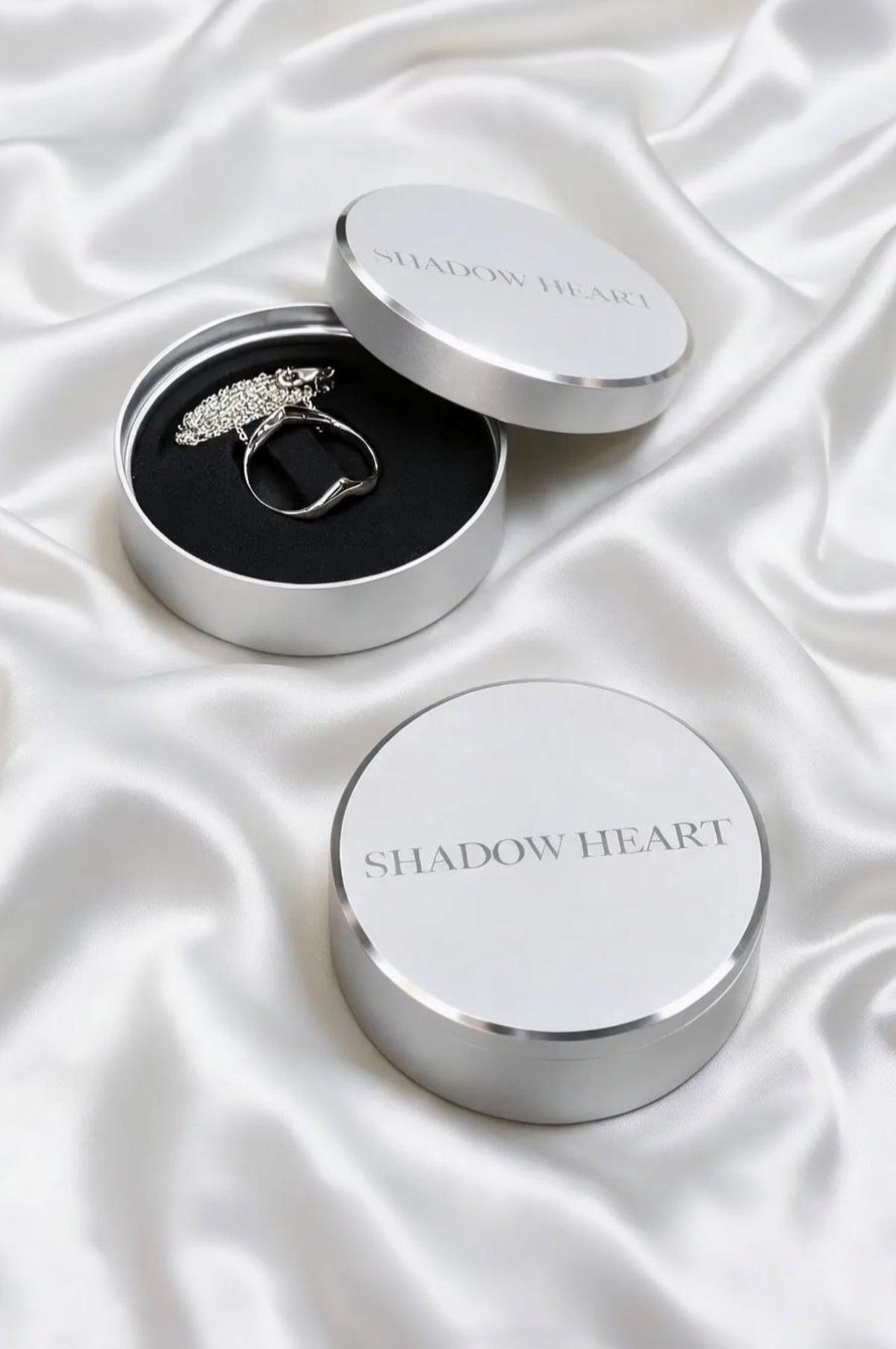A sterling silver necklace inside an open round silver box with a black foam insert. The matching lid, engraved with 'SHADOW HEART', rests beside it. A second closed box sits in the foreground on a draped white satin background.