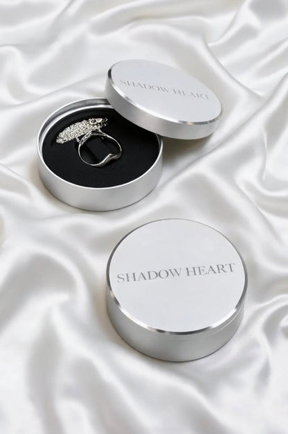 A sterling silver necklace inside an open round silver box with a black foam insert. The matching lid, engraved with 'SHADOW HEART', rests beside it. A second closed box sits in the foreground on a draped white satin background.