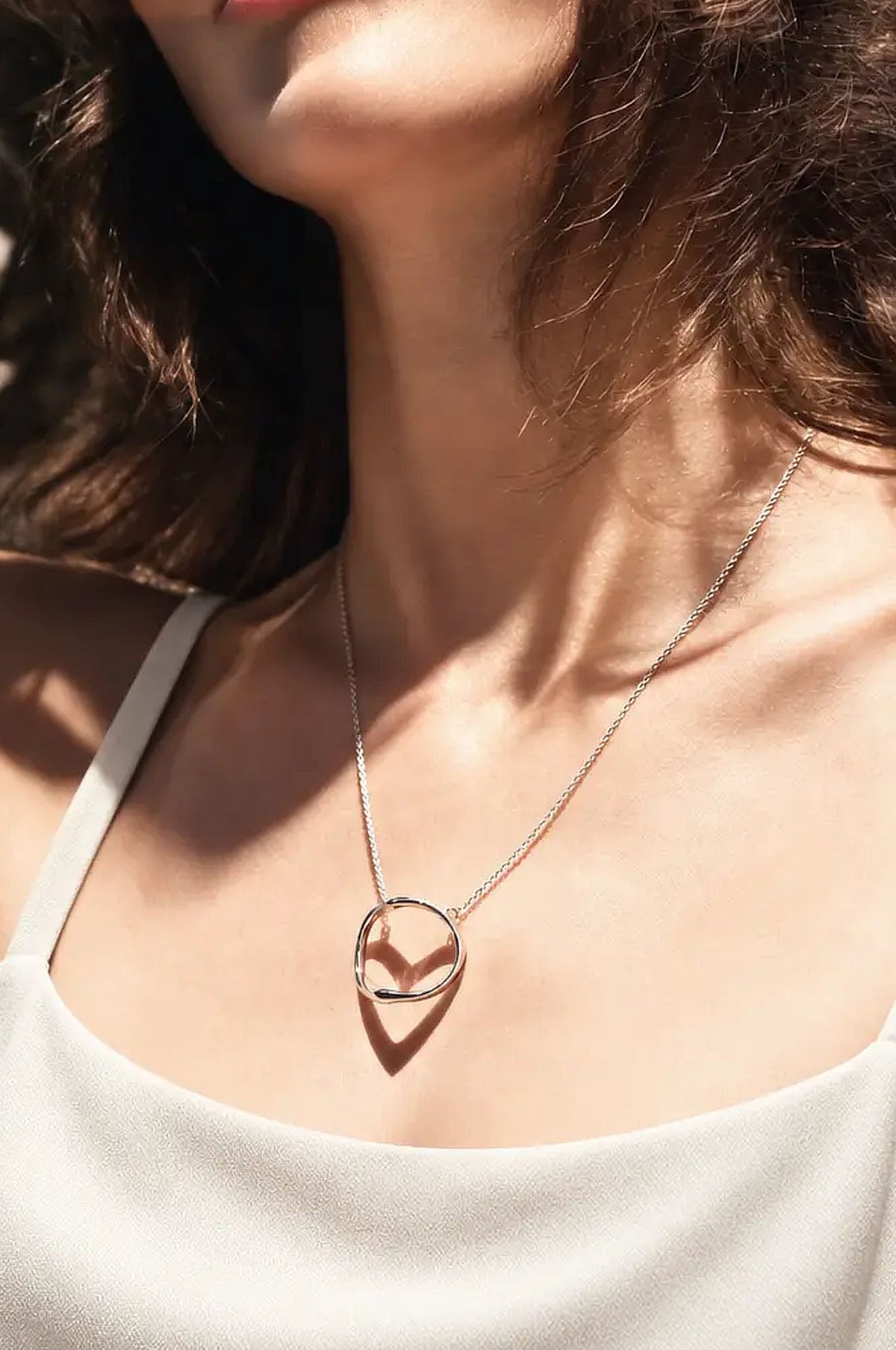 A close-up of a woman wearing a delicate silver chain necklace. The minimalist ring-shaped pendant casts a heart-shaped shadow, styled with a white camisole in warm sunlight.