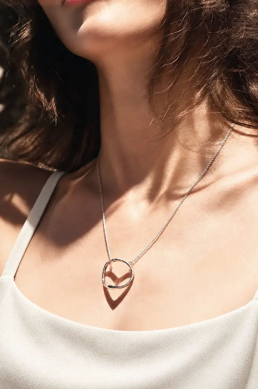 A close-up of a woman wearing a delicate silver chain necklace. The minimalist ring-shaped pendant casts a heart-shaped shadow, styled with a white camisole in warm sunlight.