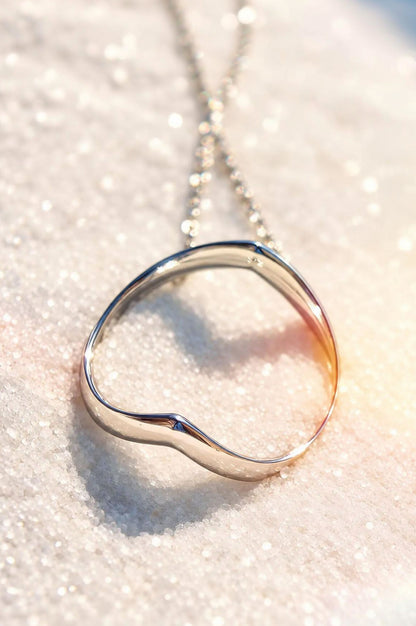 A polished sterling silver pendant with a unique design and matching chain. It rests on sparkling white sand, casting a distinct heart-shaped shadow.